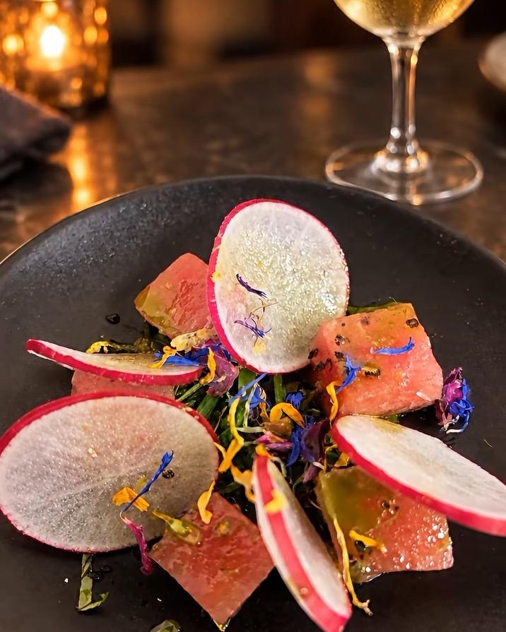 Tuna with shaved radish, basil and edible flowers on a black plate, a glass of Sauvignon Blanc behind